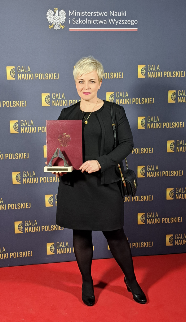 Elegancko ubrana kobieta, prof. Monika Brzychczy-Włoch, pozuję z nagrodą na tle z napisami "Gala Nauki Polskiej"